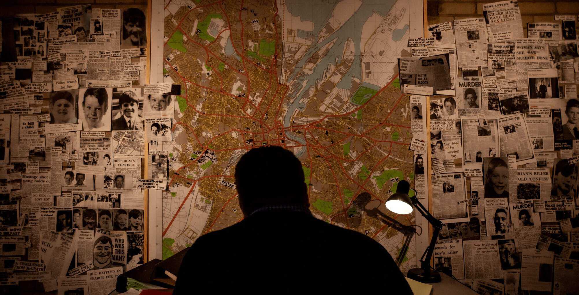 Shadow of a man set against a spotlighted map of Belfast streets with pictures and clippings from newspapers pinned to the wall in front of him