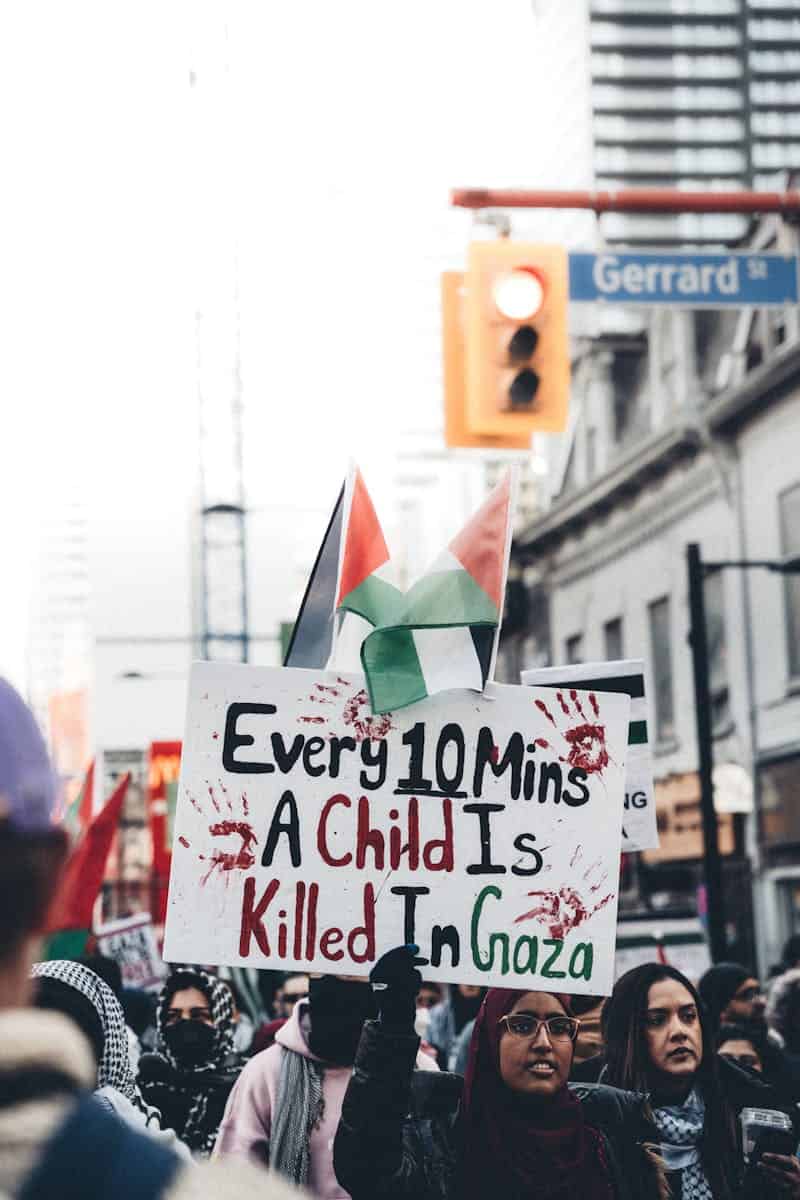 Large group of people protesting on city street with Palestinian flags and signs.