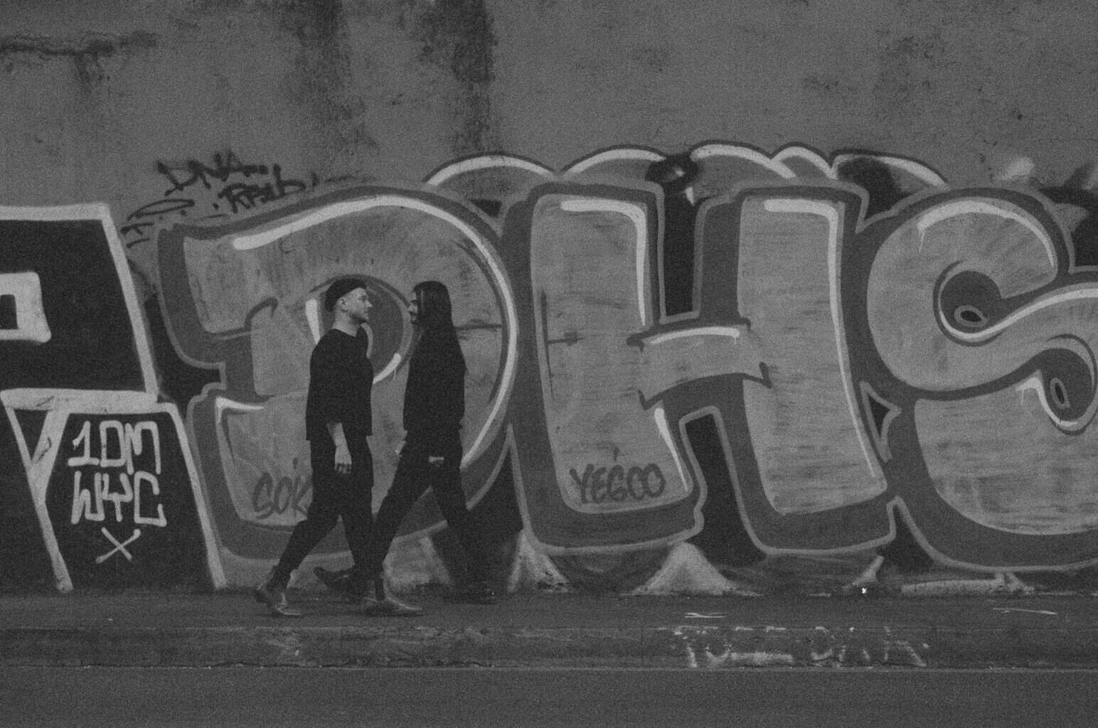 Two figures walk past graffiti on a wall.