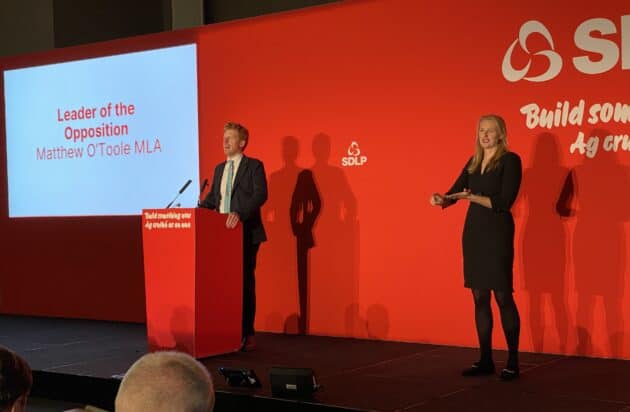 Matthew O'Toole on stage at the SDLP October 2025 conference. To one side stands sign lanuage interpreter Kristina Laverty