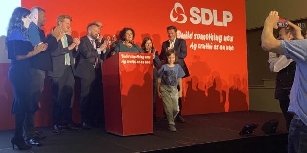 Claire Hanna surrounded by family and political colleages at the conclusion of her speech at the October 2025 conference. The conference stage backdrop is a vivid red colour.