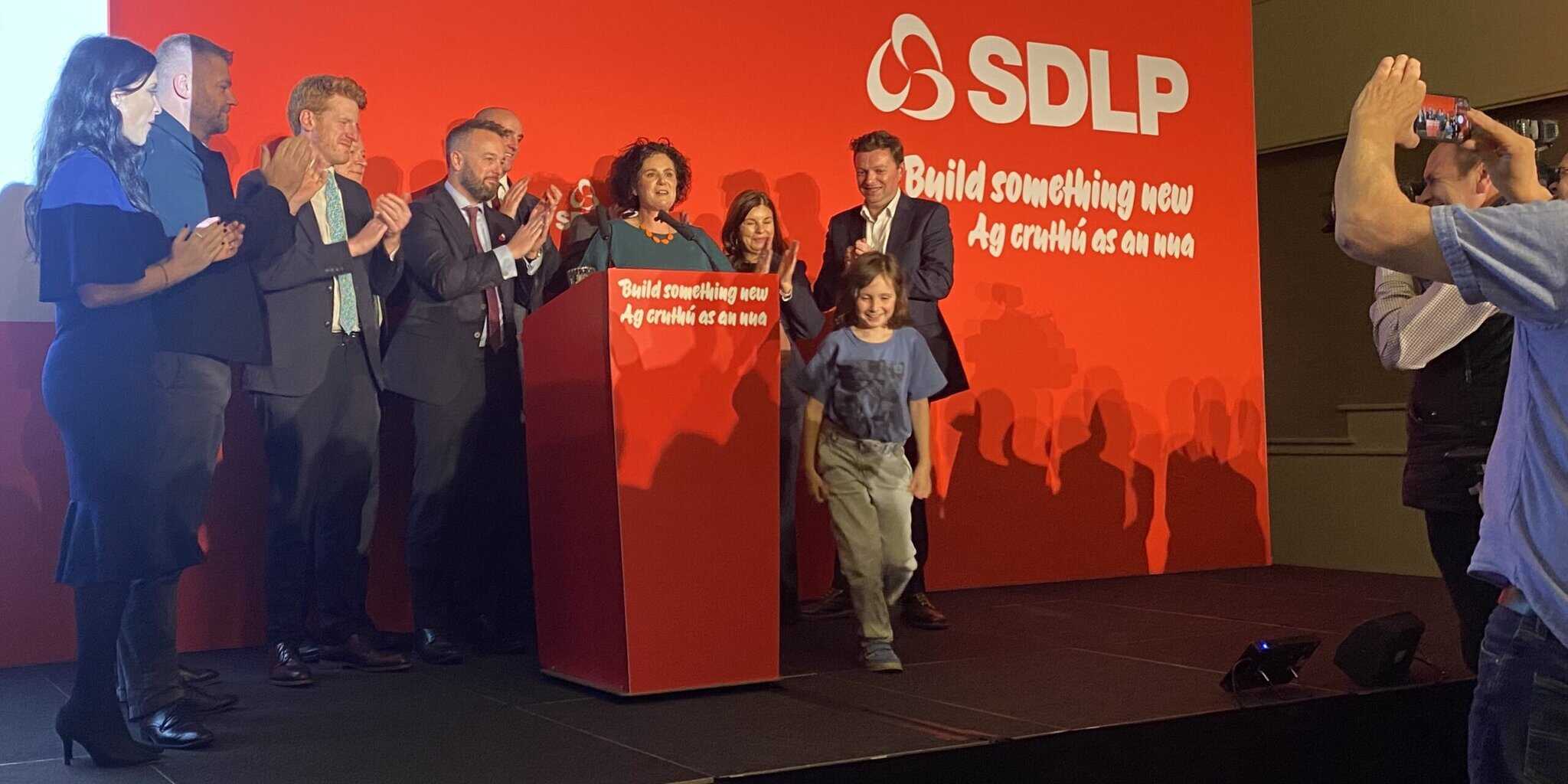 Clare Hanna surrounded by family and political colleages at the conclusion of her speech at the October 2025 conference. The conference stage backdrop is a vivid red colour.