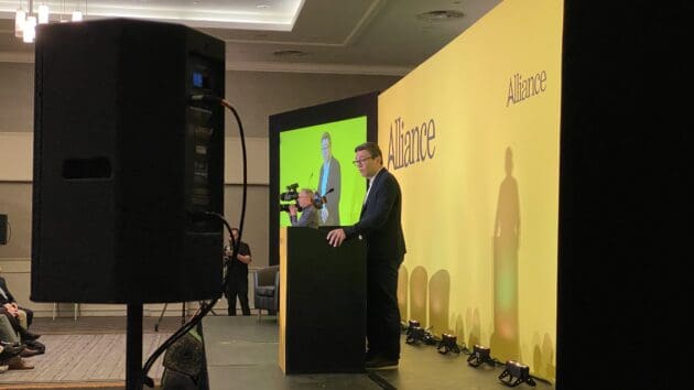 Mayor of Greater Manchester Andy Nurnham standing behind the podium on the yellow-branded Alliance Party Conference stage.