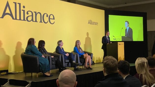 Alliance party deputy leader Eoin Tennyson speaking from behind the podium on the conference stage. Four people sit to one side of him.