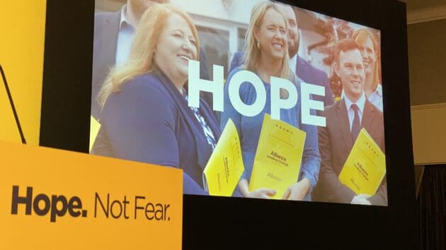 Photo of stage at Alliance Party conference. The projector image has the word HOPE overlaid on a picture of three elected reps - Naomi Long, Kate Nicholl and Eoin Tennyson. The text on the yellow podium says Hope Not Fear.