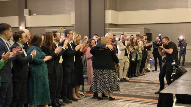 Audience standing to applaud party leader Naomi Long who is being hugged by her husband Michael.